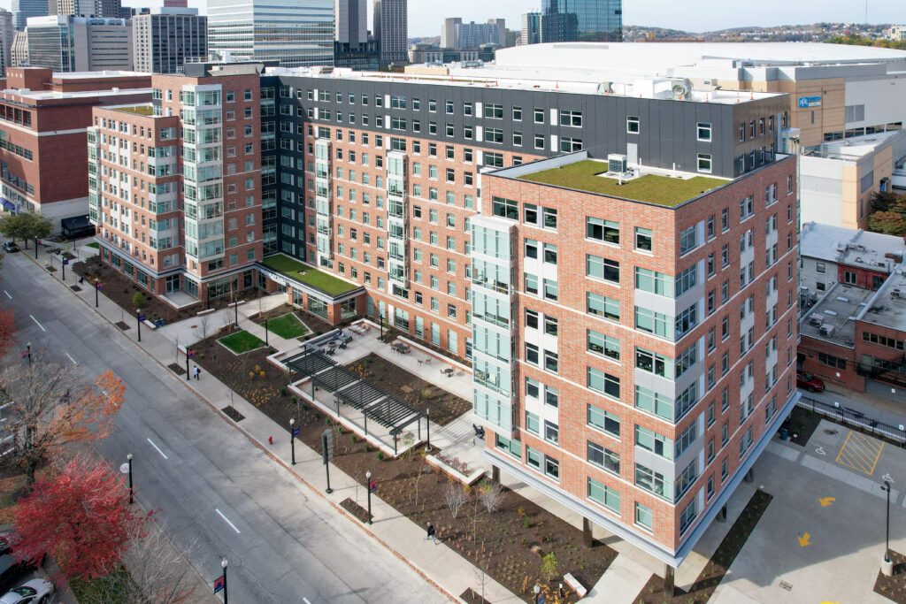 Aerial view of a multi-story brick apartment building with a rooftop garden, adjacent to a street and city skyline.