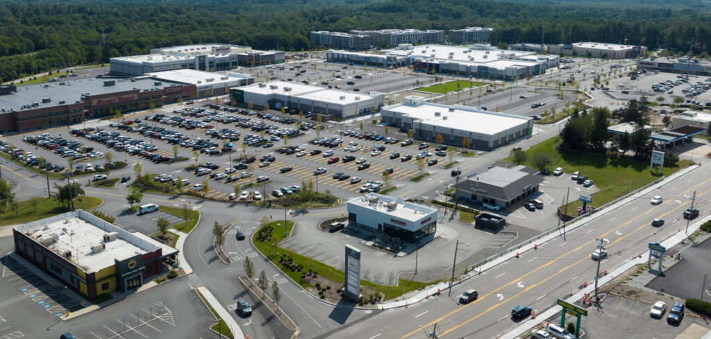Aerial view of a sprawling shopping center with large parking lots, retail buildings, and a multi-lane road in the foreground.