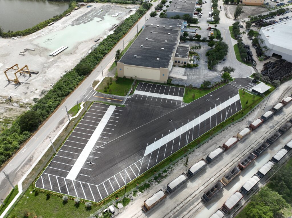 Aerial view of an industrial building next to a newly paved parking lot, with a line of freight train cars on adjacent tracks.