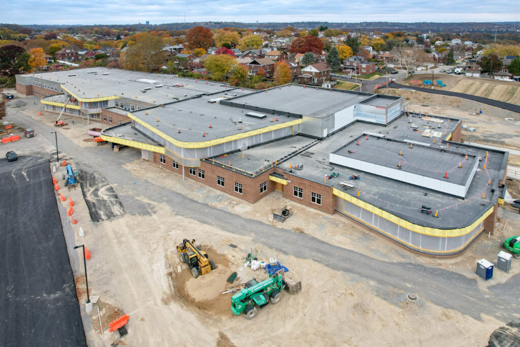 Aerial view of a school building under construction, showing a large flat roof and surrounding work site with heavy equipment.