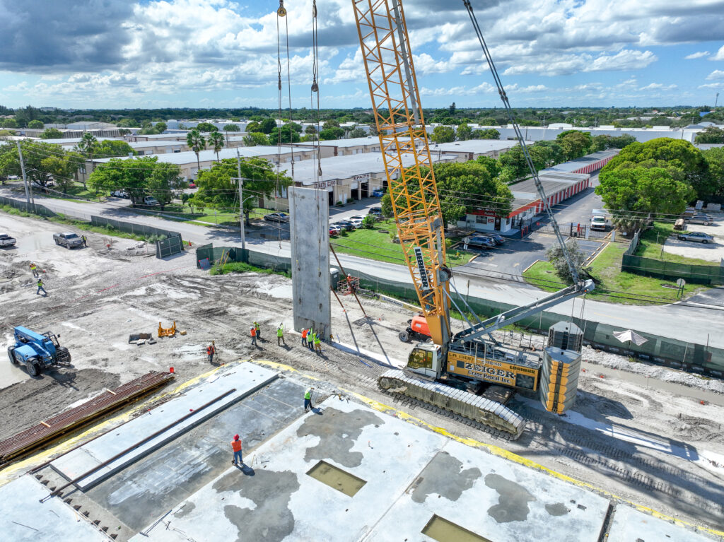A large yellow crawler crane lifts a tall concrete slab at a construction site while several workers stand nearby.