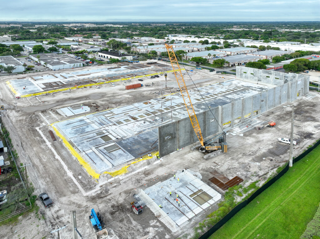 Aerial view of a construction site showing a crane lifting a concrete wall panel into place for a building foundation.