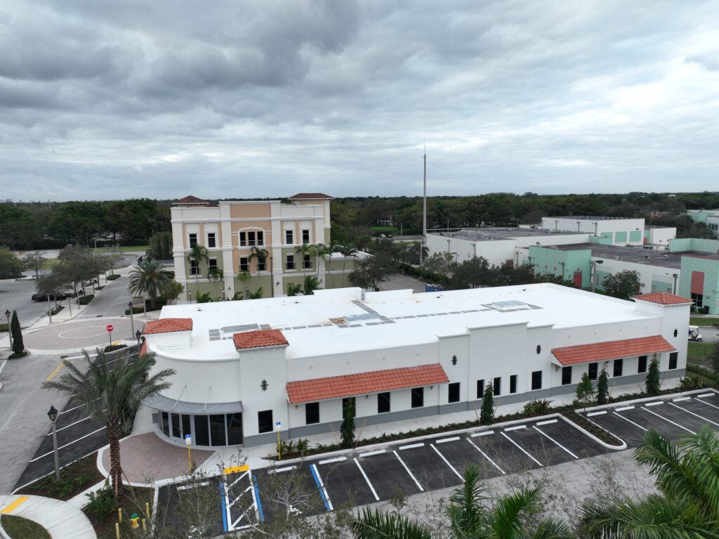 Aerial view of a white, low-rise commercial building with a terracotta roof, located in a parking lot under a cloudy sky.