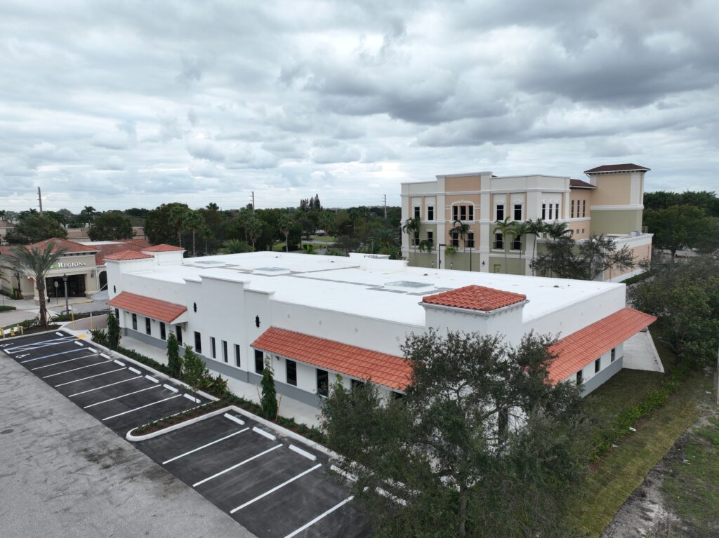 An aerial view of a white single-story commercial building with a red tile roof and an adjacent parking lot, with a taller tan office building visible in the background.