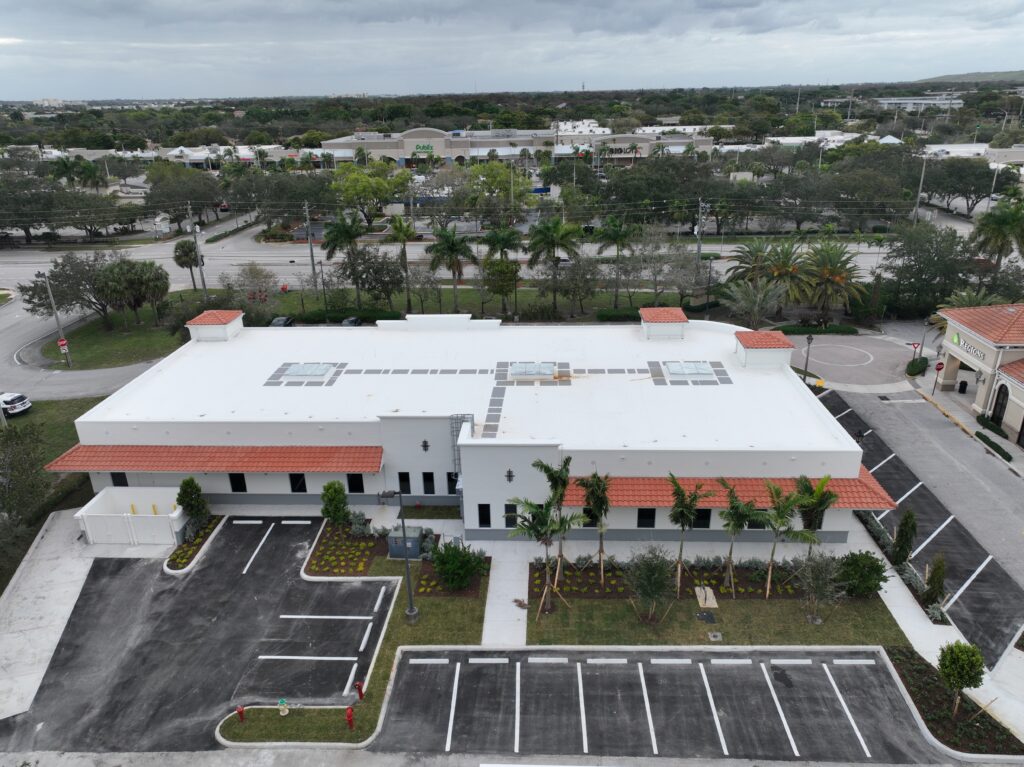 Aerial view of a white single-story commercial building with a red-tiled roof and parking lots, located across from a shopping center.