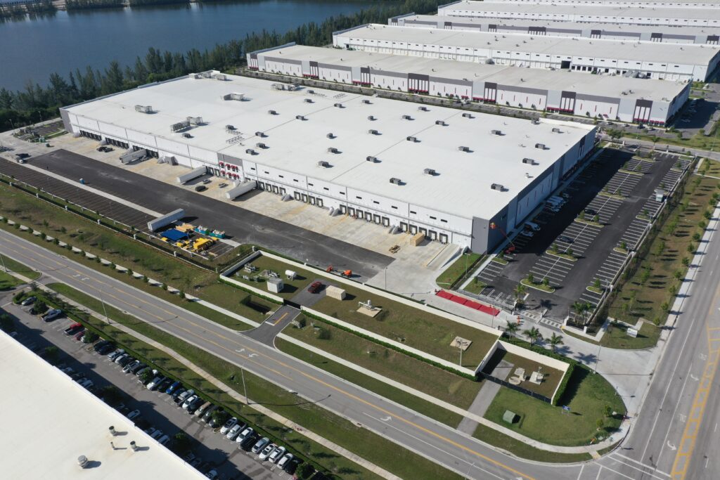 Aerial view of a large white distribution warehouse with numerous loading docks, surrounded by a parking lot and a road.