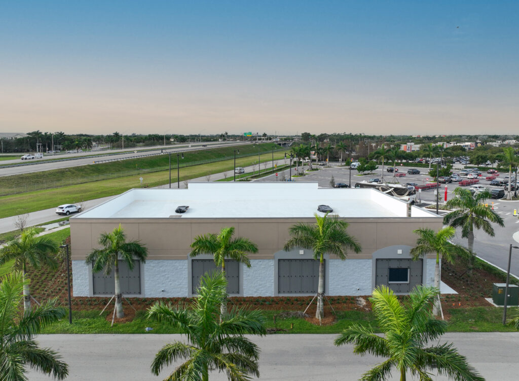 Aerial view of a commercial building with a white roof and tan walls, surrounded by palm trees, a parking lot, and a highway.