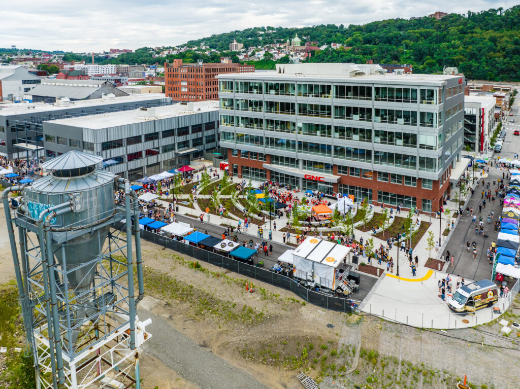 Aerial view of an outdoor community market with tents and food trucks gathered in a plaza next to a multi-story office building.