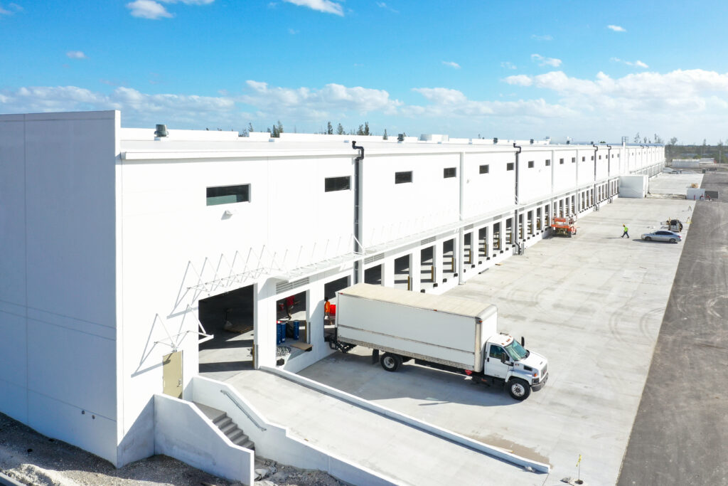 An aerial view of a long white warehouse building with multiple loading docks and a semi-truck parked at one of the bays.