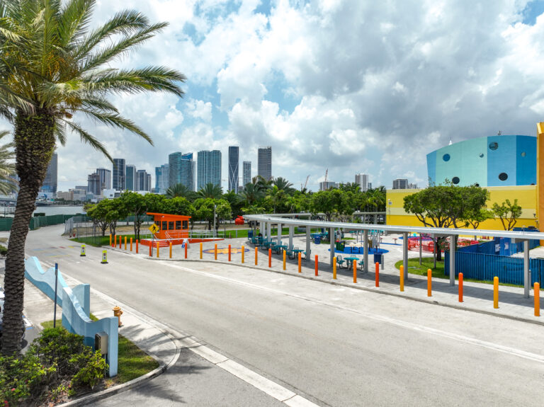 A paved road leads past an orange security booth and a covered outdoor playground area toward a city skyline under a cloudy sky.
