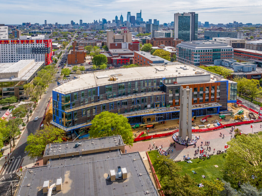 Aerial view of a campus construction site with a partially completed modern building and a tall concrete monument in a plaza, with the Philadelphia city skyline in the background.