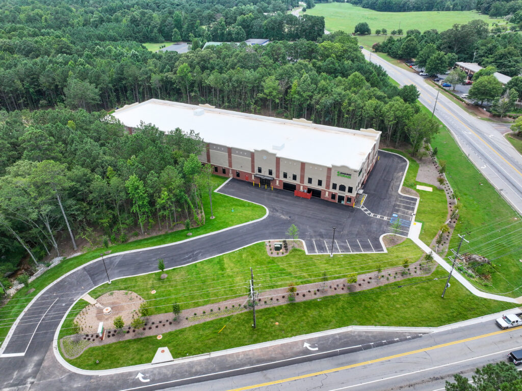Aerial view of a long, flat-roofed storage facility surrounded by trees, with an asphalt parking lot and access roads.