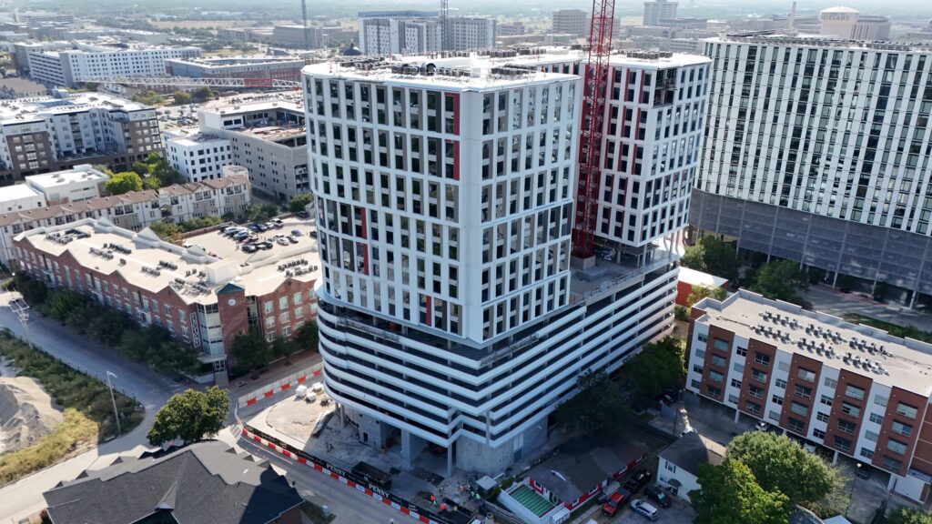 Aerial view of a large, modern high-rise building under construction, featuring a white grid facade, red accents, and an adjacent construction crane.