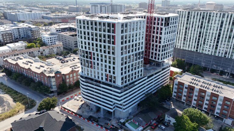 Aerial view of a large, modern high-rise building under construction, featuring a white grid facade, red accents, and an adjacent construction crane.