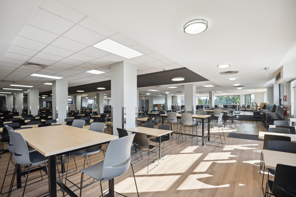 An open-plan cafeteria or workspace with rows of light wood tables and grey chairs.