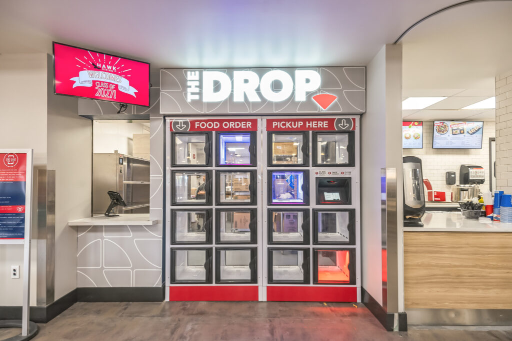A food pickup kiosk labeled “THE DROP” with rows of individual compartments, situated next to a service window and a digital menu board.
