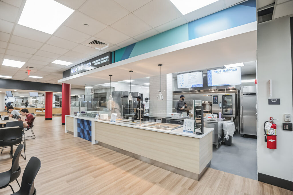 True Balance“ dining station with a serving counter, food preparation equipment, and staff members, located in a cafeteria.