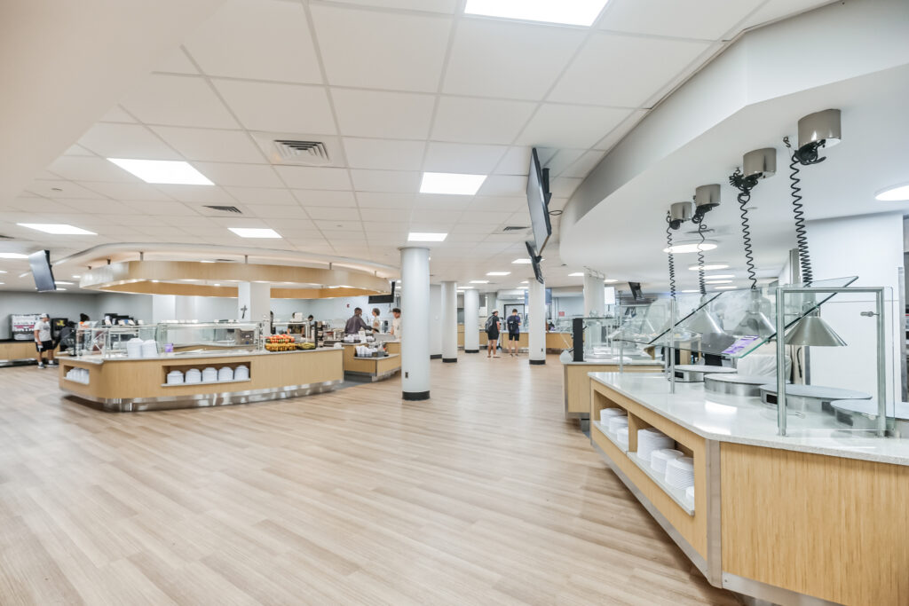 A spacious, modern dining hall with light wood counters, food serving stations, and people browsing the selection.