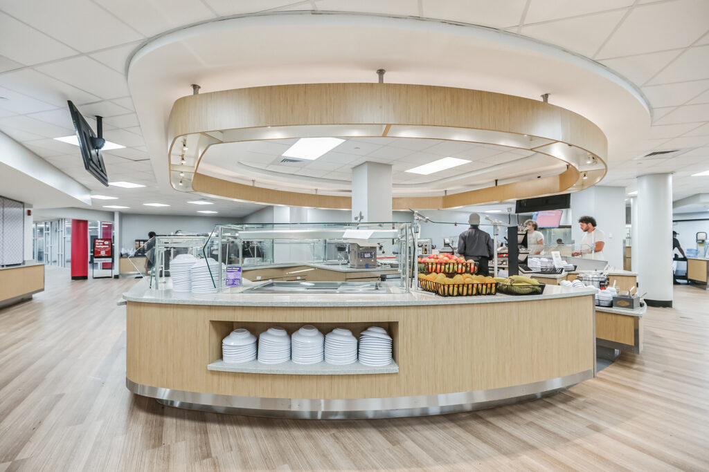 A curved cafeteria serving counter with stacked plates and bowls, fruit baskets, and staff behind a glass partition.