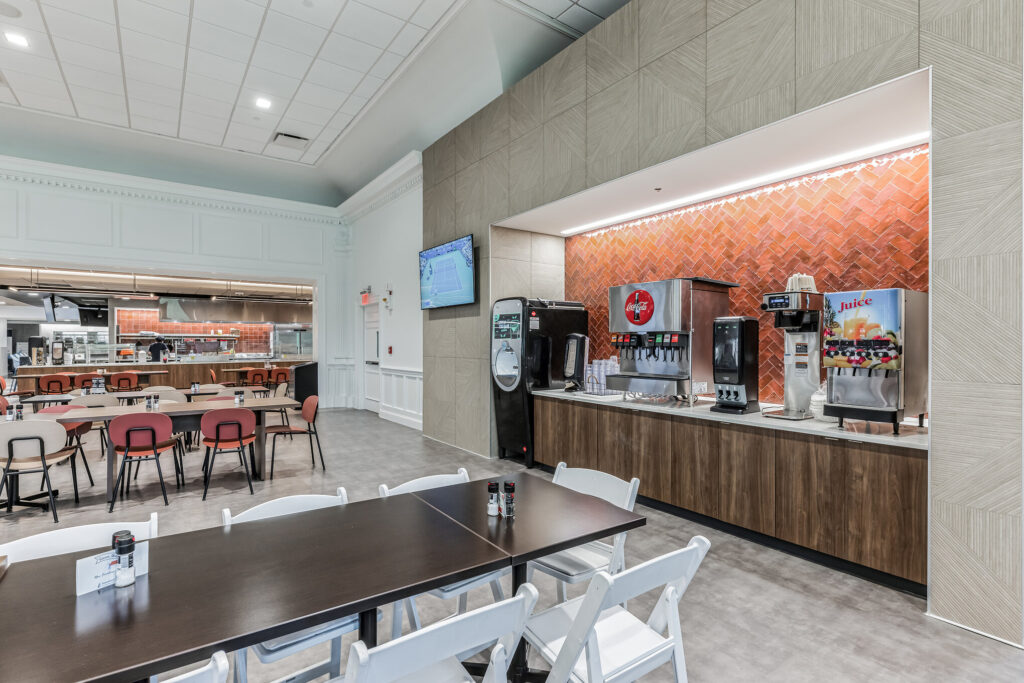 A self-service beverage station with a soda fountain and juice dispenser in a cafeteria with rows of tables and chairs.