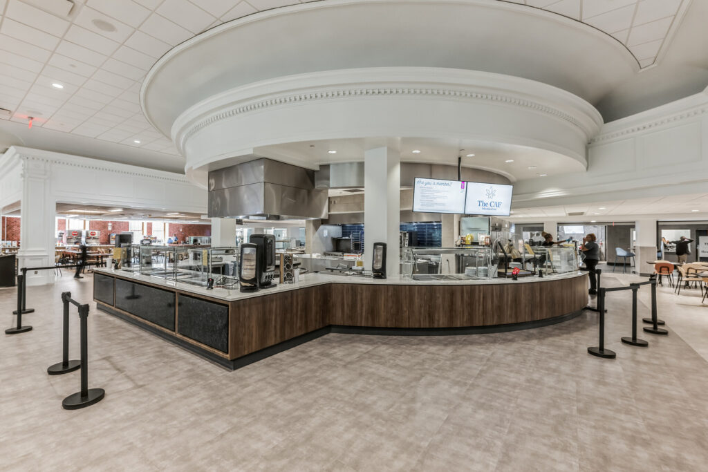 A curved cafeteria serving counter with stainless steel equipment, overhead menu screens, and stanchions for queueing.