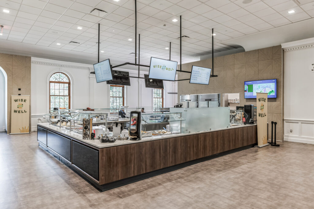 Wise Well cafeteria serving station with food display counters, digital menu boards, and branded signage.