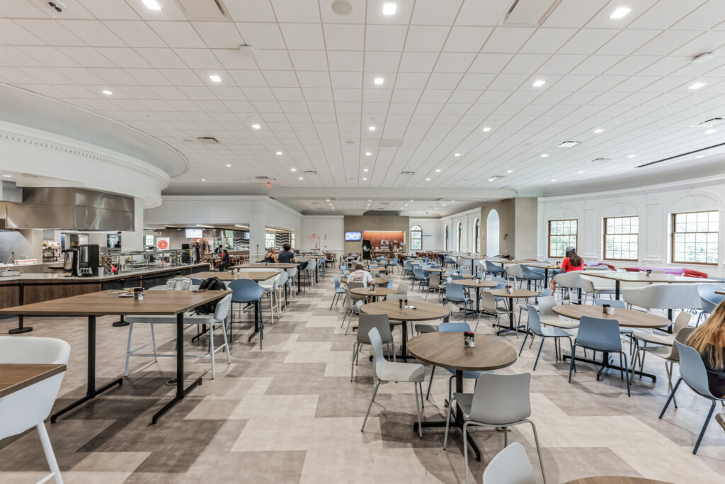 A large, modern cafeteria with rows of round and rectangular tables and chairs, including a food service counter in the background.
