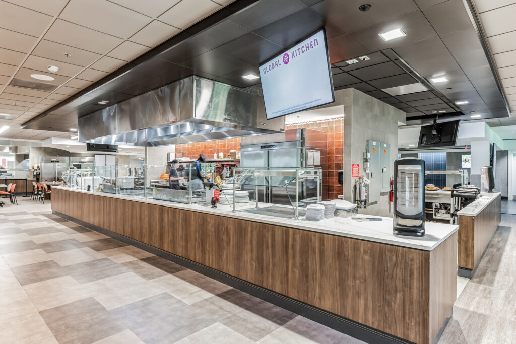 A cafeteria serving station with a stainless steel cooking area, a “Global Kitchen” overhead sign, and a long counter with stacks of plates.