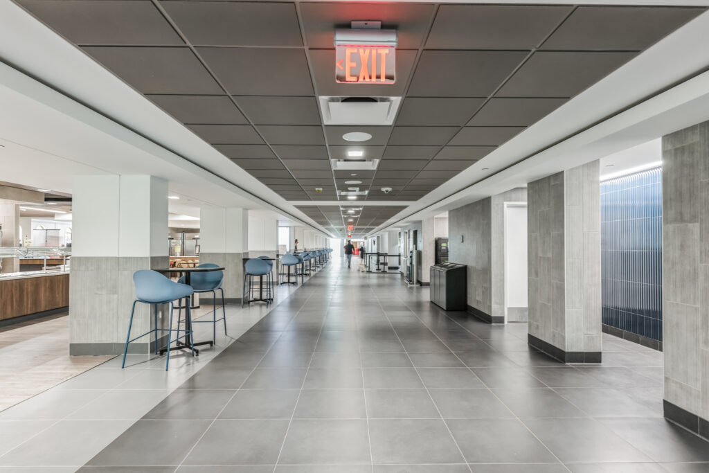 A long hallway with blue stools at high tables along the left wall and a glowing red exit sign on the ceiling.
