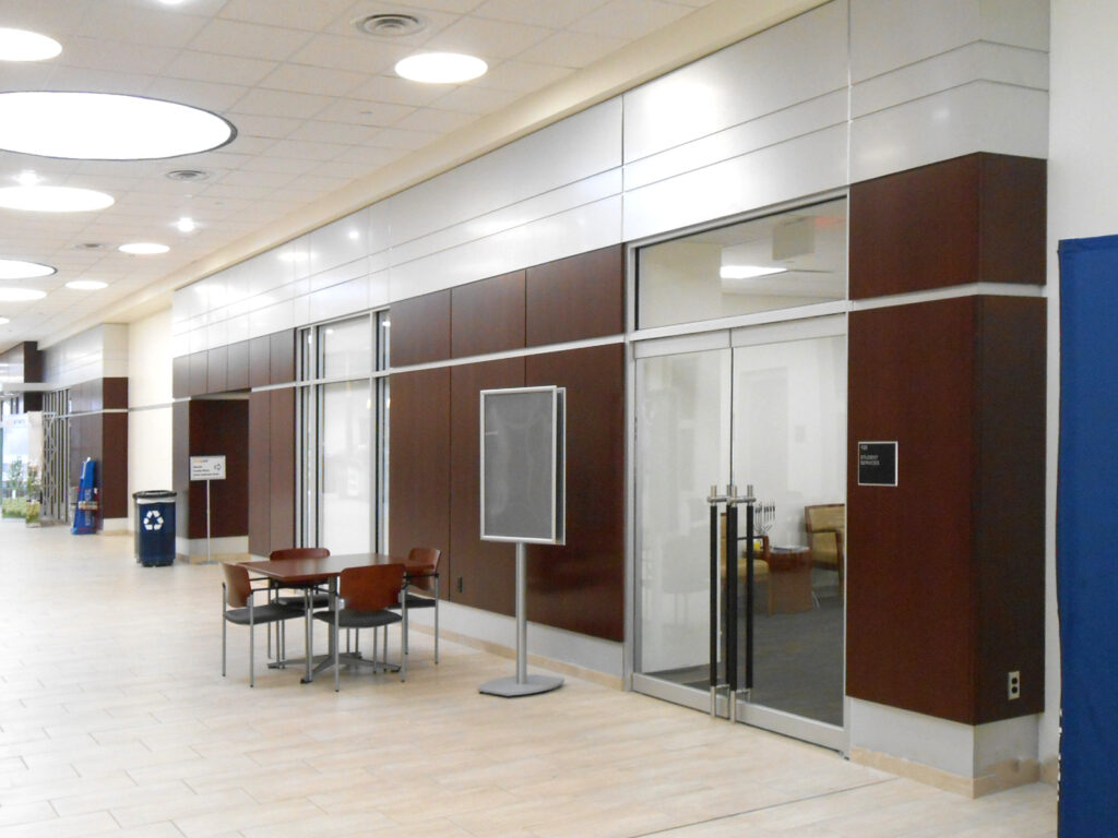 A modern office building hallway with a table and chairs, a free-standing display board, and a glass-paneled door marked “Student Services.”