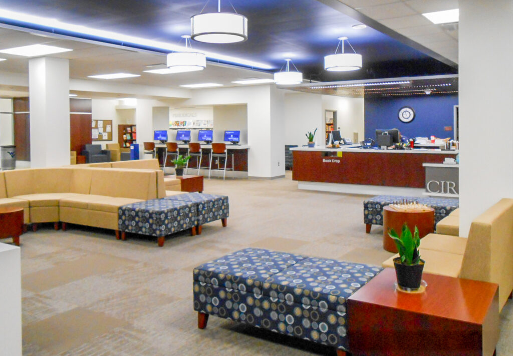 A modern library interior with beige lounge seating, patterned ottomans, a circulation desk, and a computer area with high-top chairs.