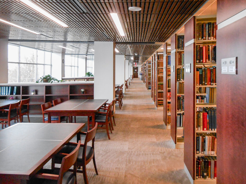 Interior view of a quiet library with long study tables in the foreground and tall, dark wood bookshelves lining the hallway.