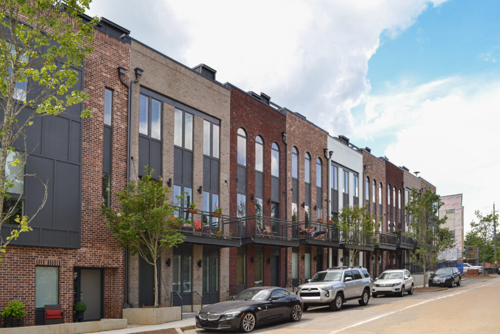 A row of modern brick townhouses with metal balconies, featuring cars parked along the street in front.