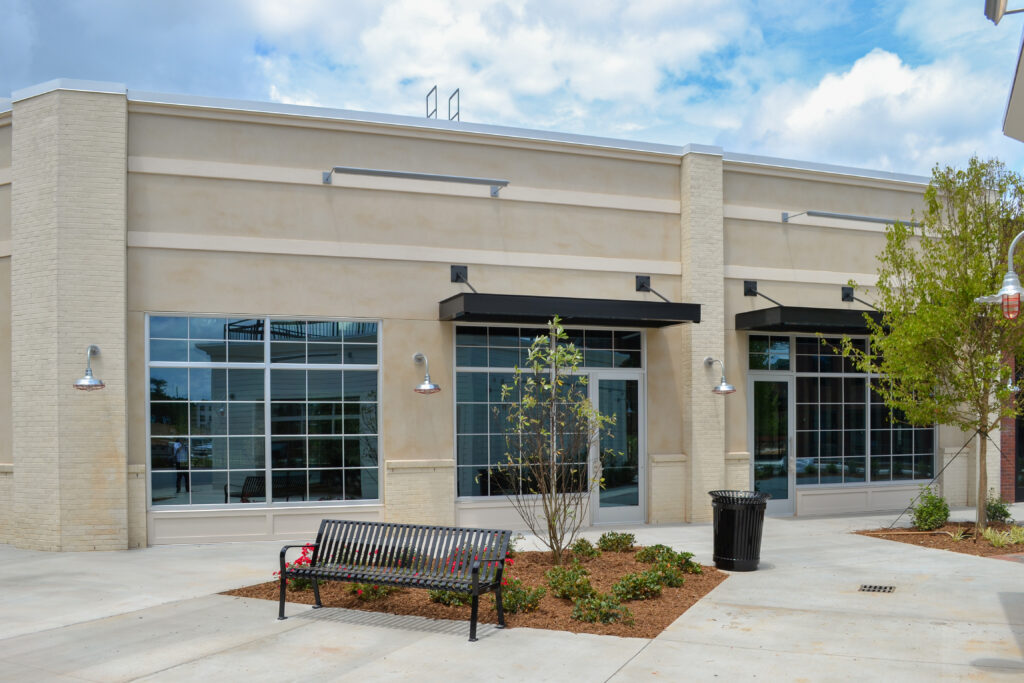 A modern beige commercial building with large glass windows, a metal bench, and a small landscaped planter in front.