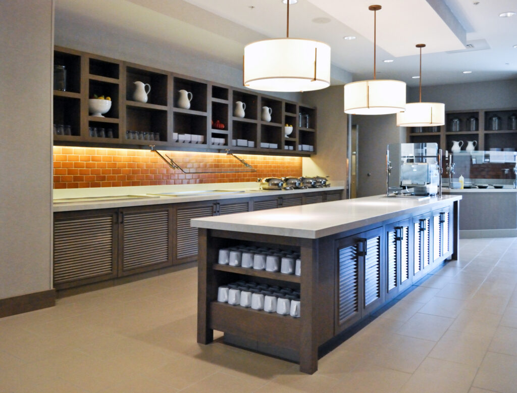 A buffet serving area featuring a long kitchen island with stacked mugs, overhead pendant lights, and shelving units filled with pitchers and serving dishes.