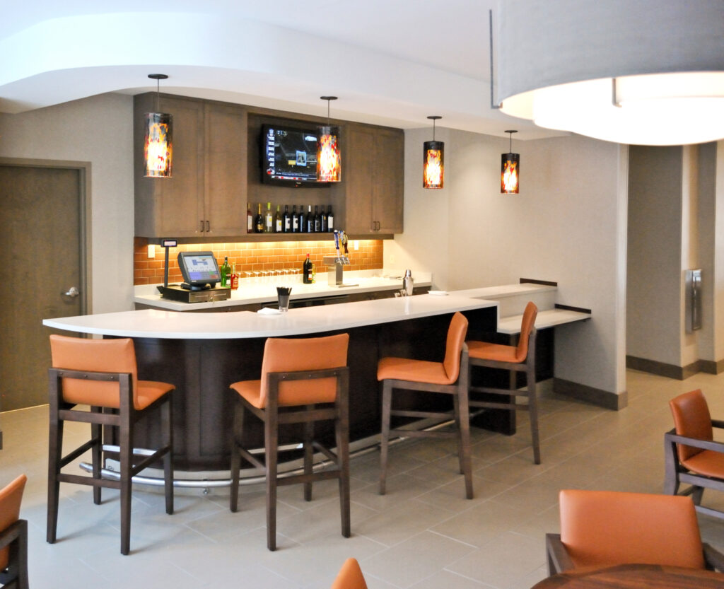 A hotel bar featuring a curved white counter, four orange bar stools, a flat-screen TV, and hanging pendant lights.