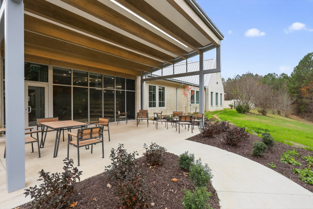 A covered concrete patio with metal chairs and tables outside a building, overlooking a grassy yard.