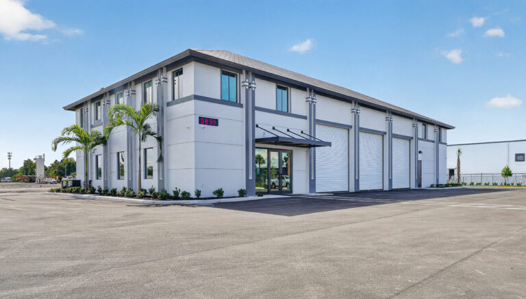 A white, single-story industrial building with three roll-up garage doors, a glass entry door, and the number 3050 displayed on the exterior wall.