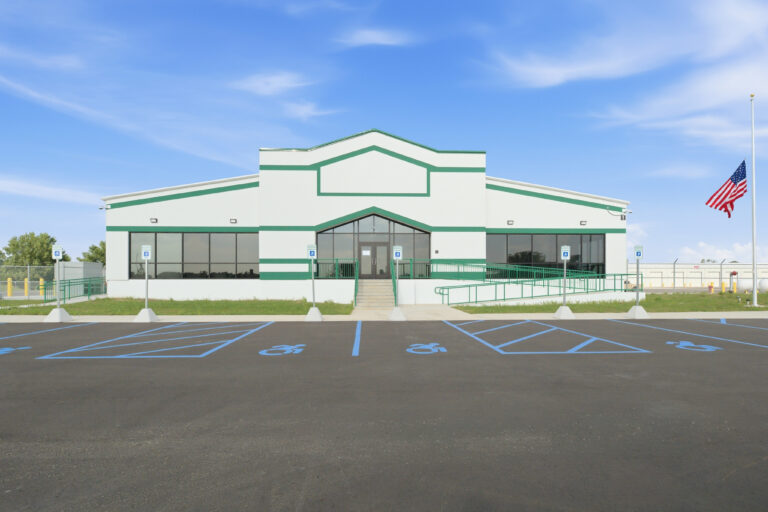 A commercial building with white walls, green trim, and an American flag on a pole, fronted by a paved parking lot with accessible spaces.