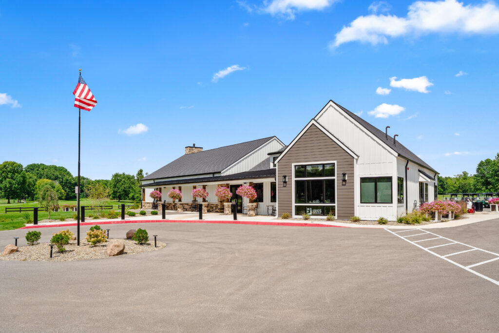 A modern clubhouse building with a dark shingled roof, white and brown siding, and hanging flower baskets, featuring an American flag on a pole in the foreground.