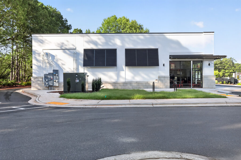 A one-story white commercial building with black window shutters and a glass entrance door, situated next to an asphalt road.