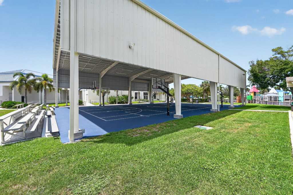 A covered outdoor basketball court with blue flooring and adjacent bleachers.