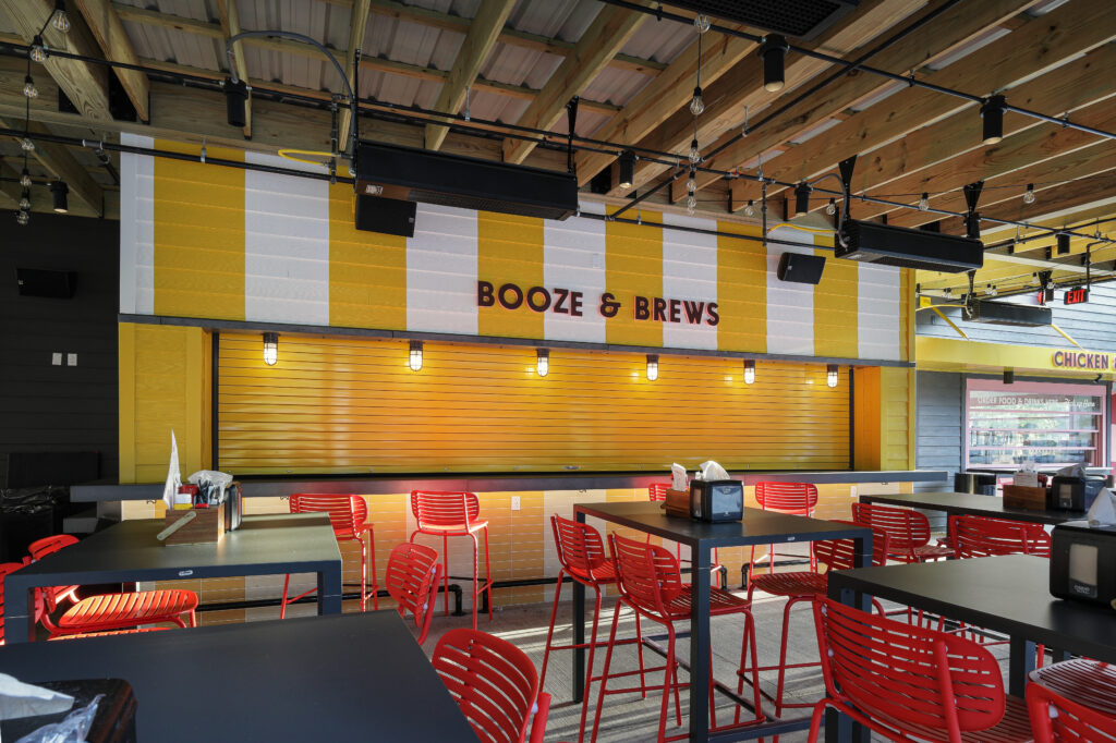 An outdoor seating area with yellow and white striped walls, red metal chairs, and a “BOOZE & BREWS” sign above a closed yellow service counter.