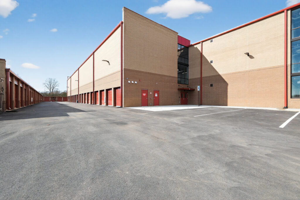 Exterior view of a multi-story storage facility with a long row of red rolling doors and a paved parking lot.