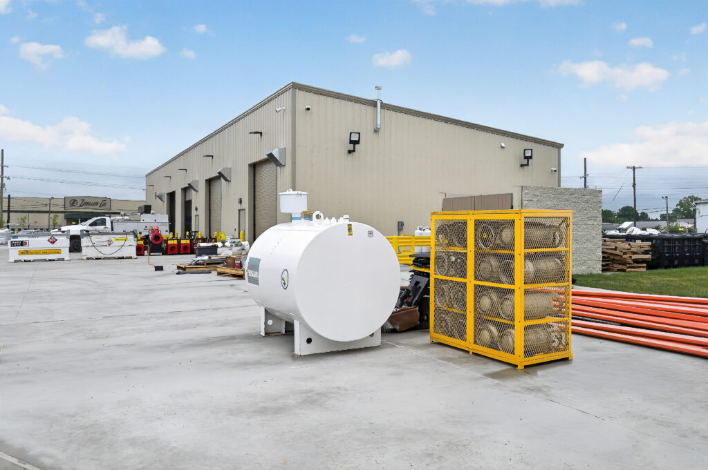 A large industrial facility exterior with a white cylindrical fuel tank and a yellow storage cage filled with gas cylinders in the parking lot.