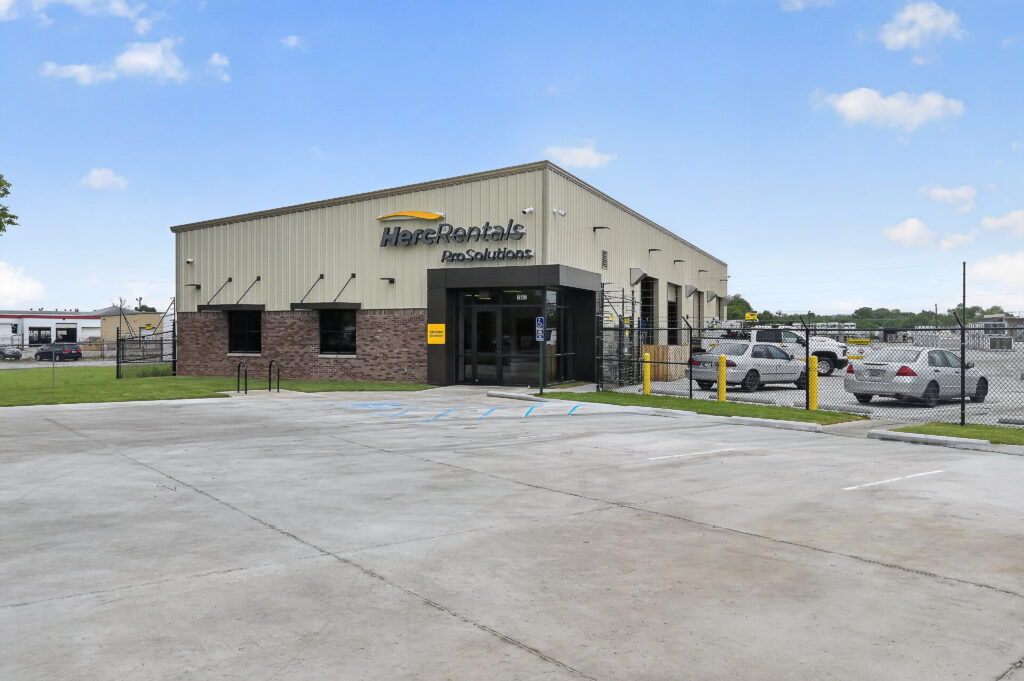 Exterior of a Herc Rentals ProSolutions building with a concrete parking lot and a fenced-in storage yard.
