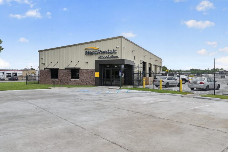 Exterior of a Herc Rentals ProSolutions building with a concrete parking lot and a fenced-in storage yard.