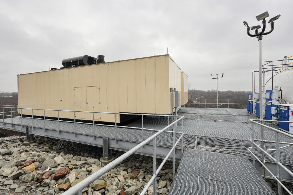 Large industrial equipment housed in tan metal enclosures on a raised steel platform, overlooking a rocky landscape under an overcast sky.