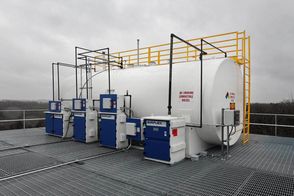 A large white industrial fuel tank with multiple Simplex control units, situated on a metal grate platform with yellow safety railings.