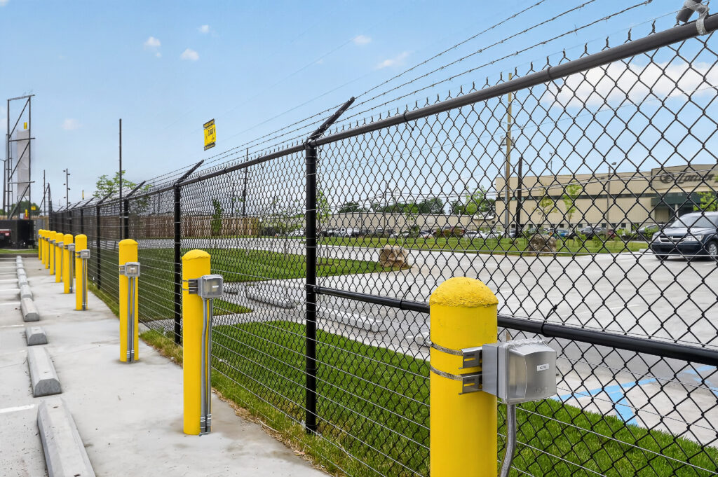 A line of yellow bollards with mounted electrical boxes positioned along a black chain-link fence topped with barbed wire.